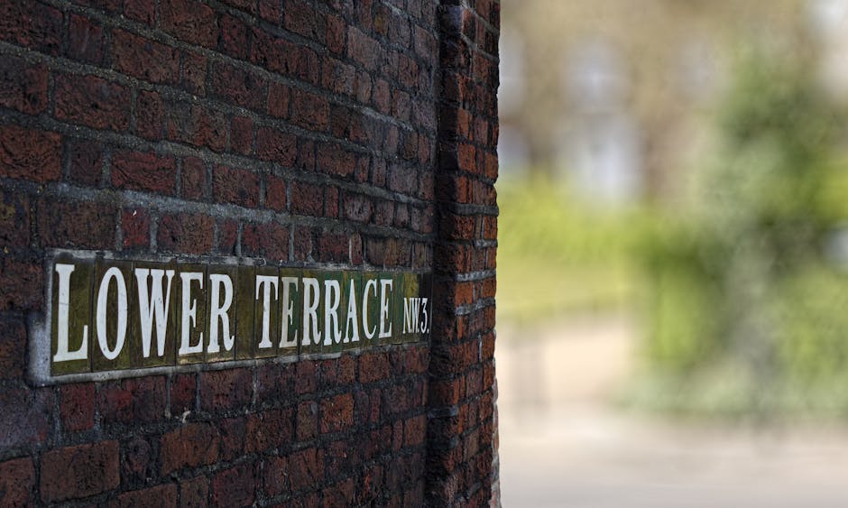A close-up view of a brick wall with a rectangular street sign that reads 'Lower Terrace N.W.3' attached to it. The sign is composed of individual ceramic tiles in shades of green, white, and black, with some tiles slightly chipped or worn. To the right of the wall, a partially open doorway reveals the interior of a building, with visible packing materials such as cardboard boxes and plastic wrapping inside. Outside, a pathway is visible with a moving trolley or dolly, likely used for furniture transport during home relocation. The background is blurred but suggests outdoor greenery and trees, indicating a residential area. The scene captures the process of packing and loading items from a property managed by Movers Hampstead during a house removal, emphasizing careful handling of furniture and packing materials in a typical urban setting.