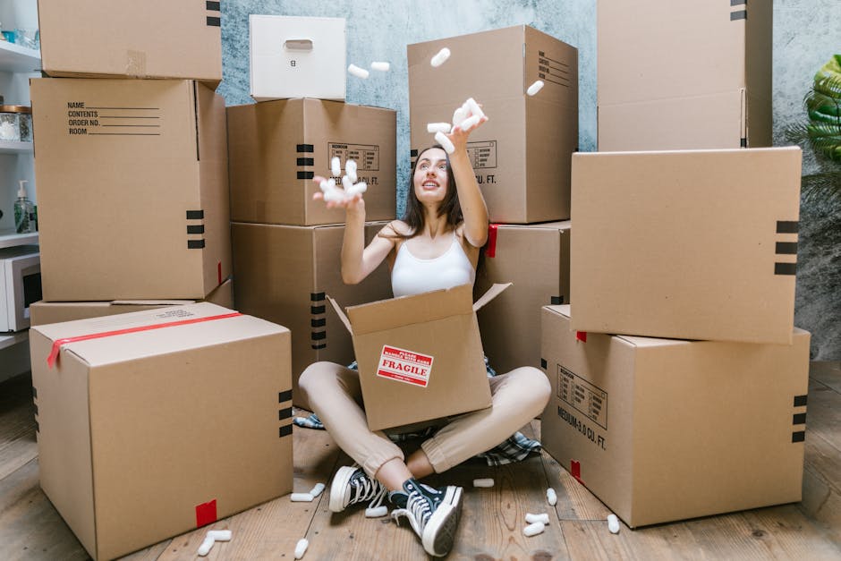 A young woman sitting cross-legged on a wooden floor inside a room, surrounded by numerous cardboard boxes of various sizes, some sealed with red tape and others open. She is smiling and appears to be in the process of packing or unpacking, with foam packing peanuts falling from her hands. The scene includes a partly open box on her lap, indicating packing activity, and the background reveals a grey textured wall with a small plant visible on the right side. This setting illustrates the packing stage of home relocation or household furniture transport, with the boxes ready for moving logistics. The environment is well-lit, emphasizing the details of the packing materials and the woman's engagement in the moving process, with a natural, professional context relevant to house removals and moving services provided by Movers Hampstead.