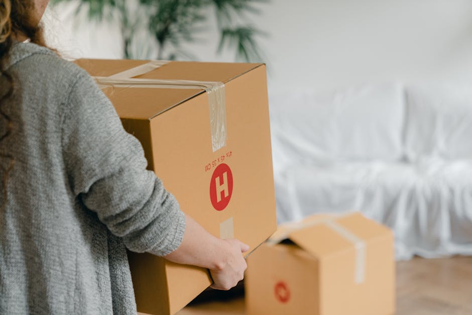 A person wearing a gray sweater is inside a property, preparing for a home relocation by taping and sealing a medium-sized cardboard box. The box has printed red markings and is being carried towards a waiting moving van or vehicle, which is visible in the background. Nearby, there is another smaller cardboard box and some plastic wrapping, indicating packing and moving preparations. The interior appears to be a living area, with a white upholstered sofa partially visible and some potted plants in the background, suggesting a residential setting. The image captures the loading process, highlighting furniture transport and packing activities carried out by movers such as Movers Hampstead as part of a house removal service in Hampstead, NW3, aligned with tips to avoid hidden fees during the relocation process.