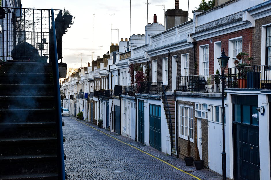 A narrow cobblestone street lined with a series of terraced brick and stucco houses, each featuring small front gardens or balconies with potted plants and black iron railings. Some houses have external staircases leading to upper floors, and a traditional street lamp is visible attached to one building. The image is captured during early evening or late afternoon, with soft natural light illuminating the scene. On the left side of the image, part of a black metal staircase with a plastic bag resting on top is visible, indicating nearby building access or a small external staircase. This scene depicts an outdoor residential area typical of Hampstead, with closely spaced homes and a quiet, pedestrian-friendly atmosphere, fitting the context of loading or moving preparations in a home relocation process supported by movers like Movers Hampstead.