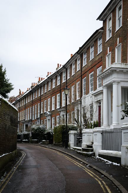 A row of traditional brick terraced houses on a narrow, slightly curved street in Hampstead, with each property featuring white-painted columns and decorative architectural details around the entrance. The street is lined with black wrought-iron fences, small front gardens with shrubs and leafless trees, and lampposts positioned along the pavement. The scene appears to be during winter, as there is a light dusting of snow on the ground and on the rooftops. A moving process is underway, with cardboard boxes wrapped in plastic and fabric moving blankets visible near the entrances, suggesting preparations for a home relocation. Movers Hampstead, a professional removal company, is likely involved in coordinating the loading and transport of furniture and packing materials, which are visible in the vicinity of the houses. The overall atmosphere is calm, with clear ambient lighting and no visible disruption, emphasizing the careful handling typical of professional house removals in urban residential settings.