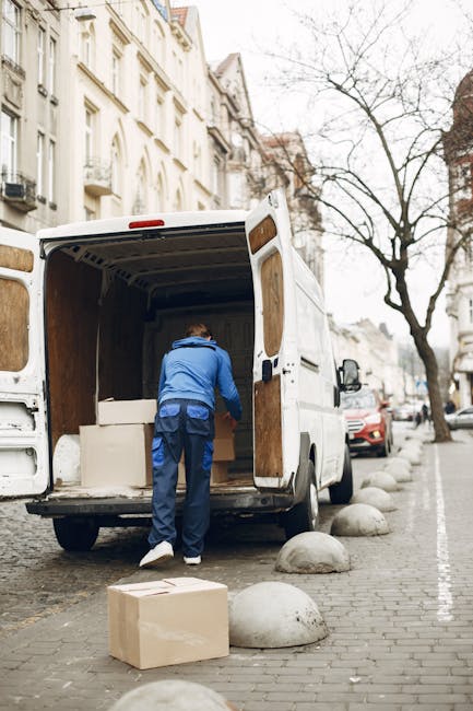 A man wearing a beige knit beanie, a grey sleeveless puffer vest over a dark blue jacket, and dark trousers stands next to the open sliding door of a black moving van. Inside the van, there are several cardboard boxes, some sealed with packing tape, stacked on the floor. The man is holding a pair of gloves in his hands and appears to be preparing to load or unload items as part of a home relocation or furniture transport process. The scene is outdoors with a paved surface, and the environment suggests a professional removals service such as Movers Hampstead, specialising in same-day house removals in Hampstead Heath.