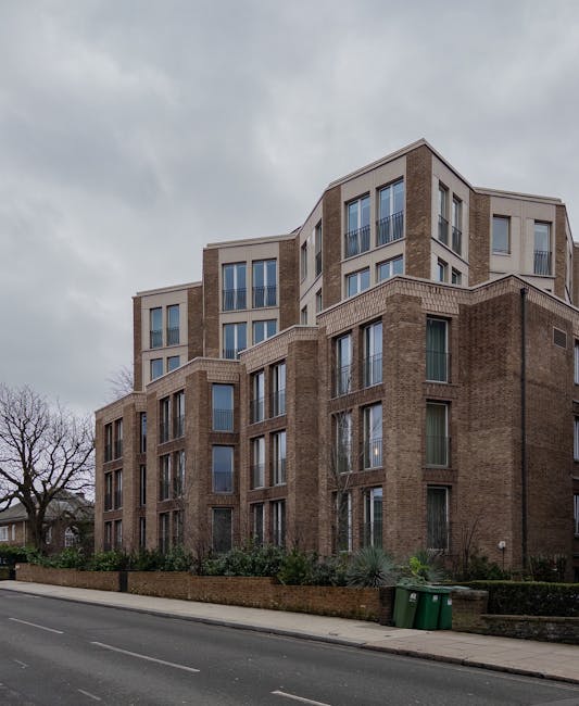 A multi-storey residential building with a modern brick facade and large rectangular windows, situated on a street in Hampstead. In the foreground, there is a paved pavement with two green waste bins positioned near the curb. The building features a combination of brown brickwork and light-colored architectural accents, with some windows having balconies enclosed by glass or metal railings. The overall scene is captured during cloudy weather, with an overcast sky above. This image relates to house removals, showing the exterior environment where Movers Hampstead might assist with home relocations, furniture transport, or packing and moving preparations inside or outside such a property.