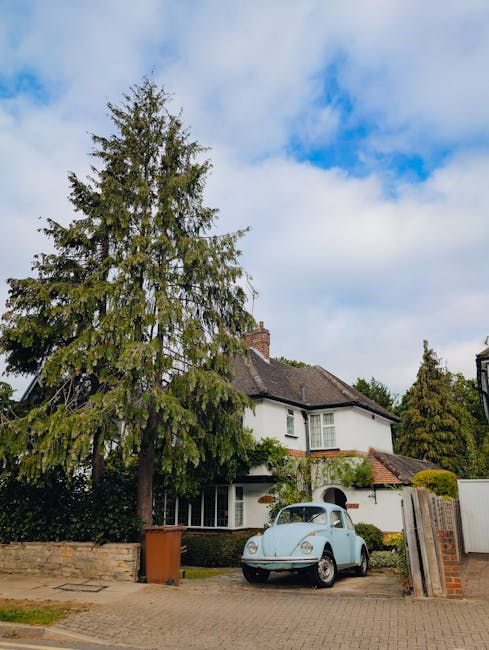 A residential property with a white, two-storey house featuring a tiled roof and multiple windows, situated behind a large evergreen tree that partially obscures the front facade. In the driveway, a vintage light blue Volkswagen Beetle is parked, with attention directed towards the area where movers from Movers Hampstead are loading furniture and boxes for house relocation. The driveway is paved with concrete and bordered by a low brick wall and wooden fencing, with a parking permit and packing materials likely nearby. The scene is captured during daytime with partly cloudy skies and natural daylight illuminating the property and moving activity, reflecting a typical home removal process involving careful furniture transport and packing support.